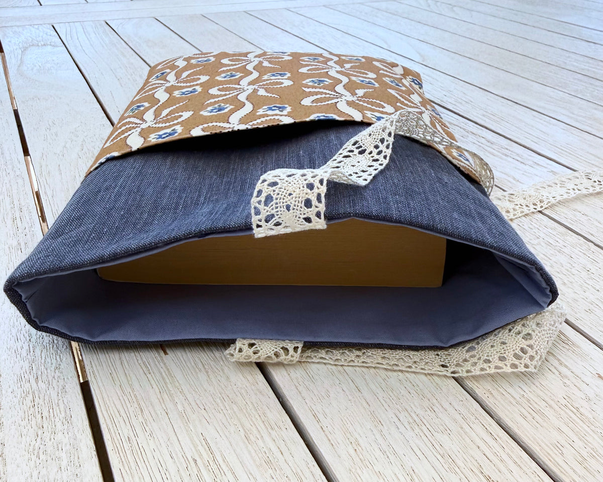 Side view of a handmade navy fabric book sleeve with a paperback book inside, showing the sleeve's opening, patterned front pocket and cream lace ribbon closure.