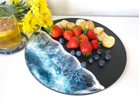 Handmade ocean resin charcuterie tray with slate base, styled with crackers, strawberries and blueberries, flowers in the background, coastal-inspired serving and cheese board for entertaining