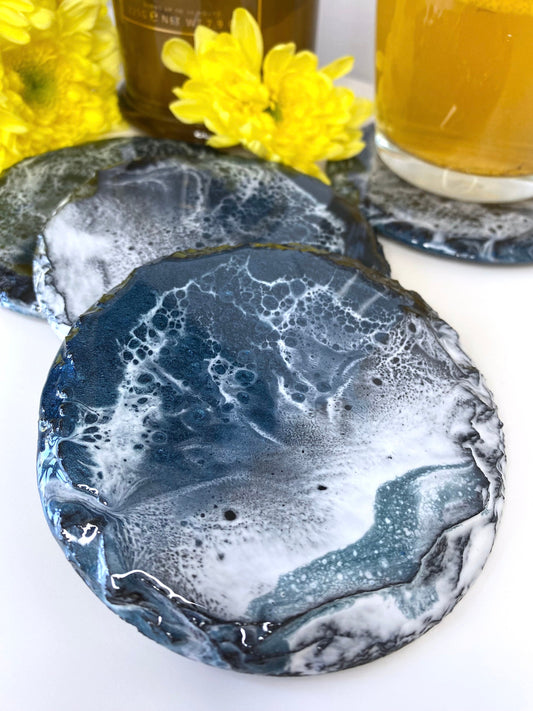Set of four round resin art slate coasters, ocean-inspired design, close-up showing waves pattern on one coaster, handmade decorative coasters for home or gifts.