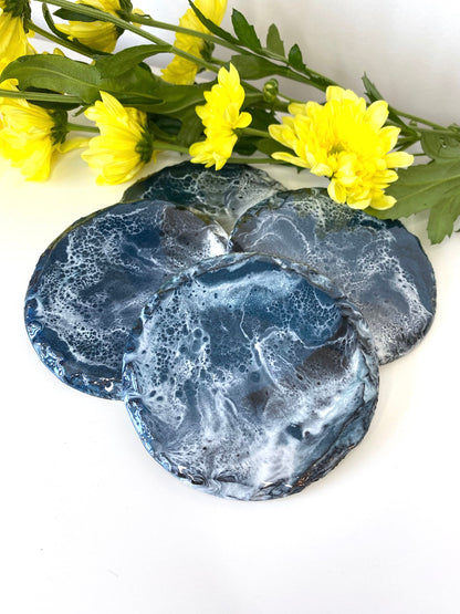 Set of four round resin art slate coasters, ocean-inspired design, scattered on table, showing individual wave patterns, handmade decorative coasters for home or gifts