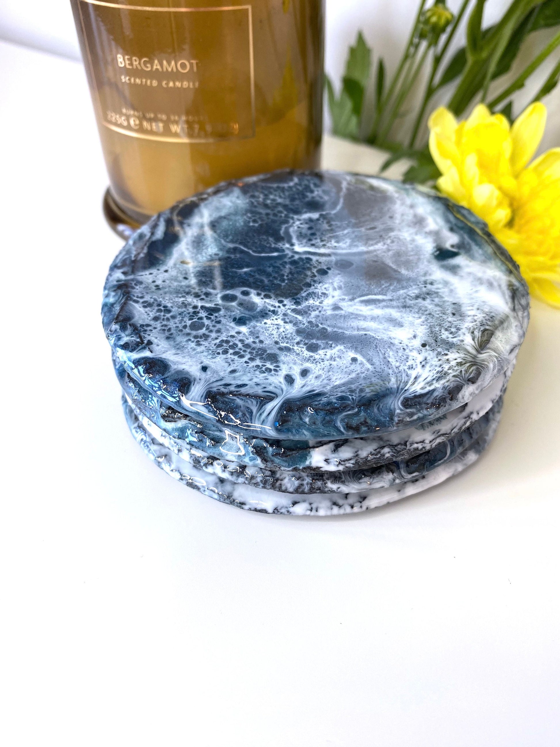 Set of four round resin art slate coasters, ocean-inspired design, stacked on top of each other on table with candles and flowers in background, handmade decorative coasters for home or gifts