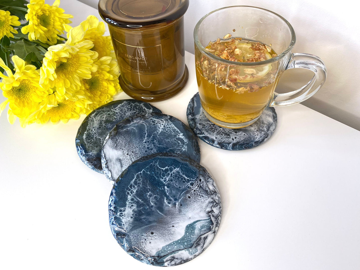 Set of four round resin art slate coasters, ocean-inspired design, styled on table with tea cup on one coaster, candles and flowers around, alternate angle showing full design on one coaster and others partially visible, handmade decorative coasters for home or gifts.