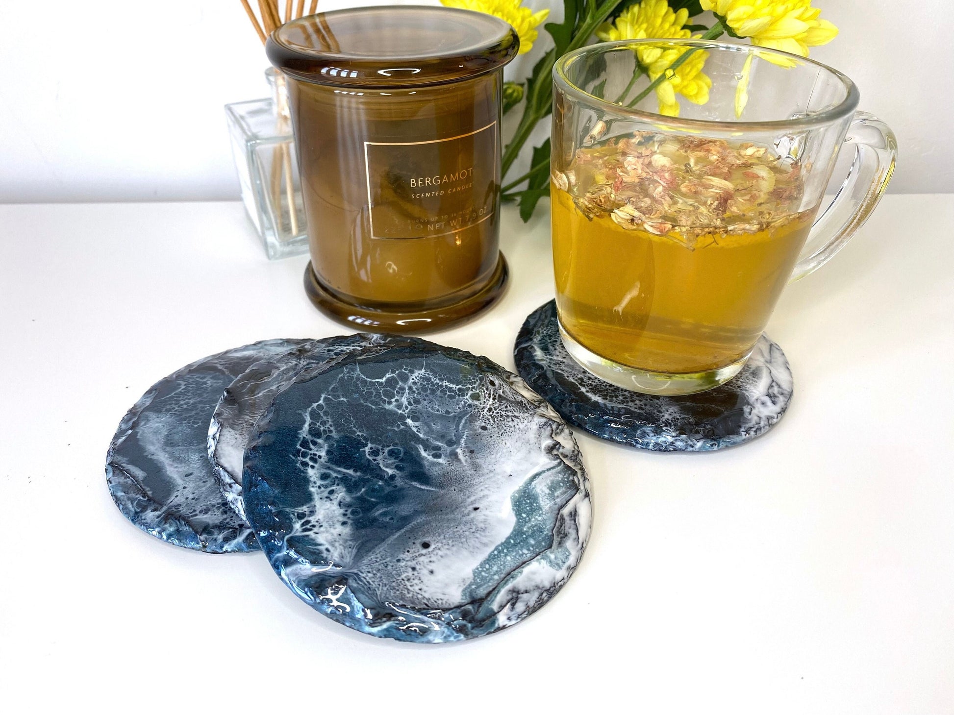 Set of four round resin art slate coasters, ocean-inspired design, styled on table with tea cup on one coaster, candles and flowers around, showing full design on one coaster and others partially visible, handmade decorative coasters for home or gifts