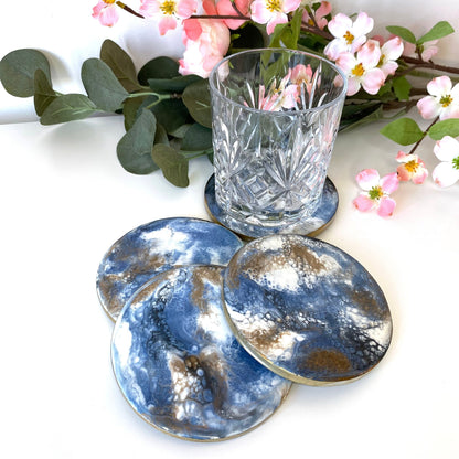 Set of 4 round slate resin coasters with blue, white, black, and gold epoxy design and gold-painted edges, three stacked domino-style with a water glass on the fourth.
