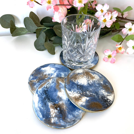 Set of 4 round slate resin coasters with blue, white, black, and gold epoxy design and gold-painted edges, three stacked domino-style with a water glass on the fourth.