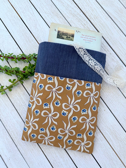 Front view of a navy denim-look paperback book sleeve with a book protruding from the top and a caramel-brown pocket decorated with cream bows and blue flowers.