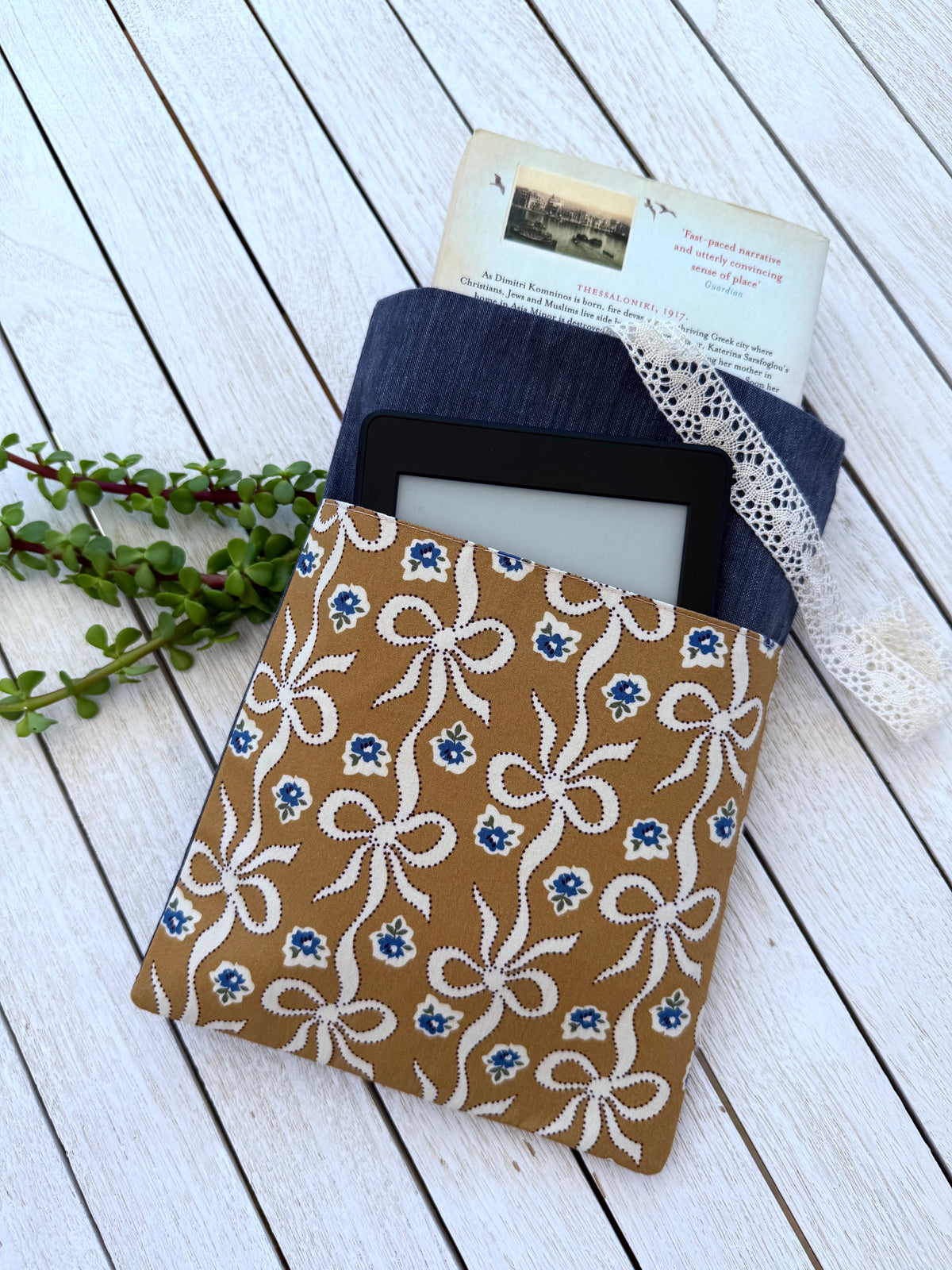 Handmade navy denim-look book sleeve holding a paperback book, with a black e-reader tucked into the caramel-brown bow print front pocket, showing how it stores reading essentials.