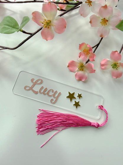 Personalised acrylic bookmark with rose gold glitter name and stars design, lying flat on a white surface with a matching tassel.