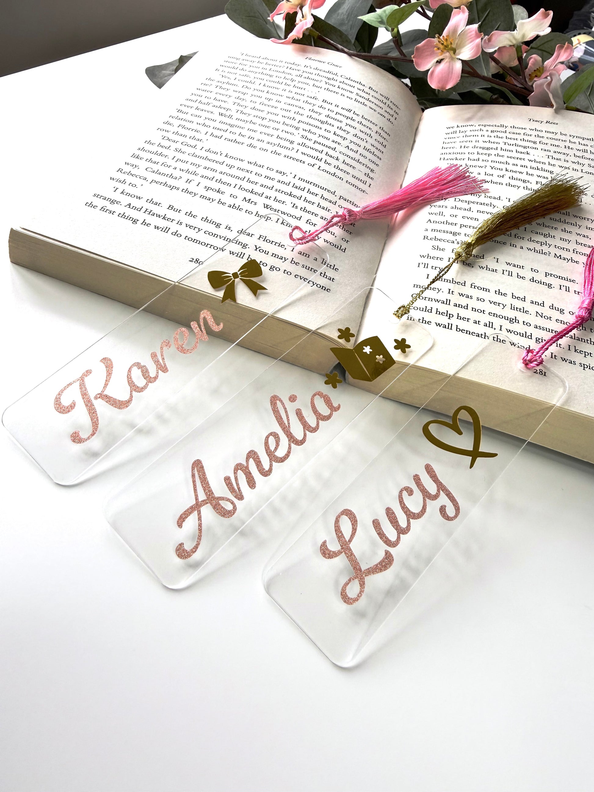 Three personalised acrylic bookmarks in rose gold glitter with bow, heart, and book designs, displayed on an open book with the bottoms resting on a white surface and flowers in the background.