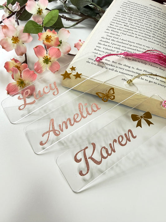 Personalised acrylic bookmarks in rose gold glitter with stars, butterfly, and bow designs, displayed on an open book with flowers in the background.