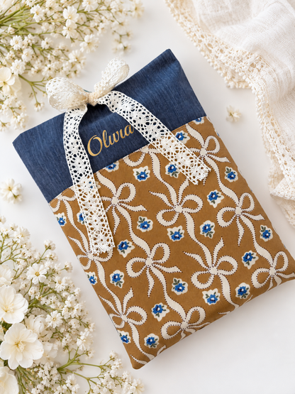 Personalised navy denim-look book sleeve with a gold vinyl name, caramel-brown bow print pocket and cream lace ribbon closure, styled with white flowers.
