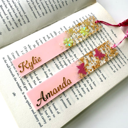 Two personalised pink resin bookmarks on an open book: one with butterfly design, one with floral design, both customised with gold vinyl and matching tassels.
