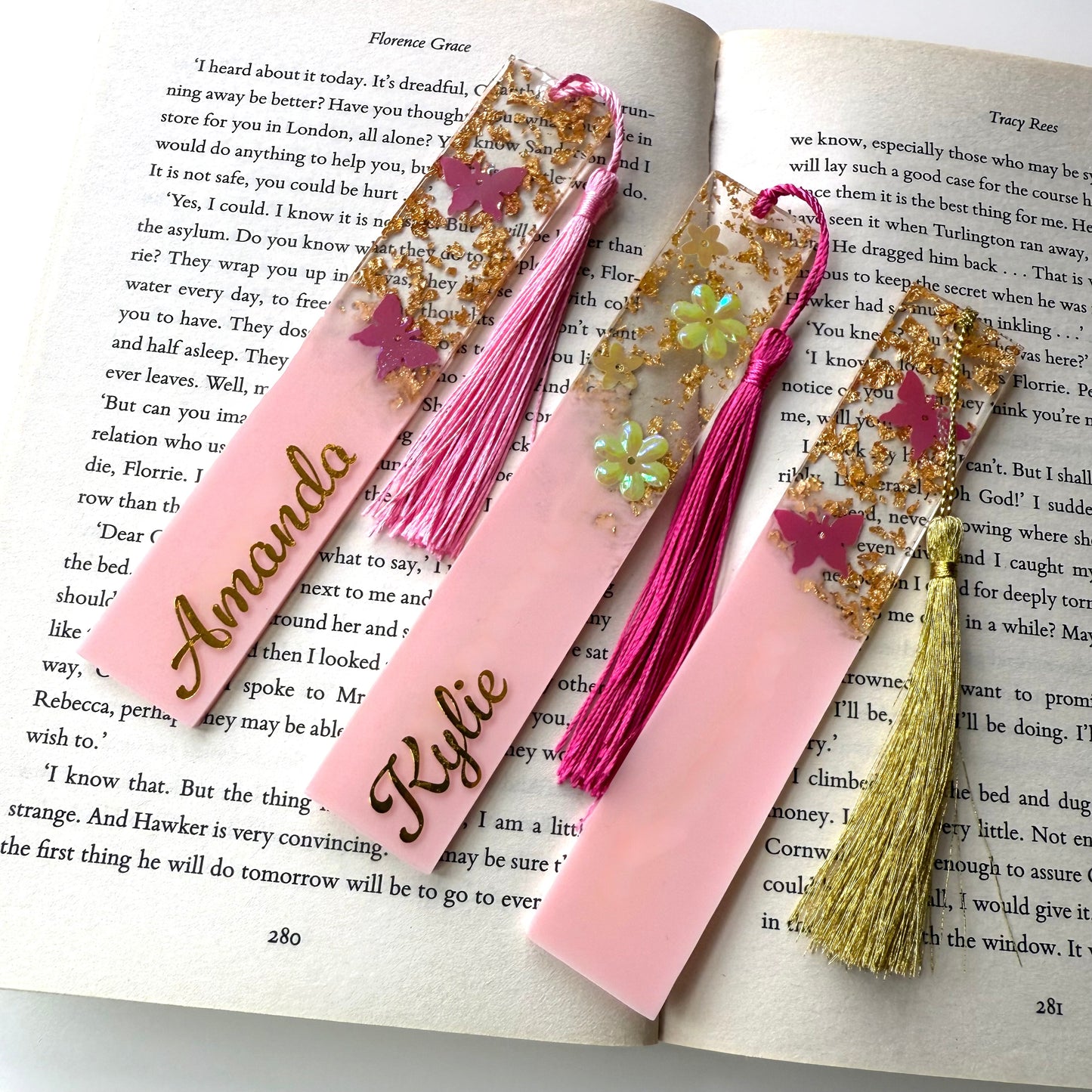 Three handmade resin bookmarks on an open book: personalised pink butterfly bookmark, personalised pink flower bookmark, and non-personalised pink butterfly bookmark, each with a matching tassel.