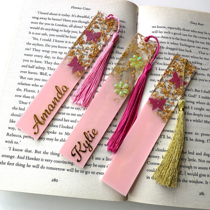 Three handmade resin bookmarks on an open book: personalised pink butterfly bookmark, personalised pink flower bookmark, and non-personalised pink butterfly bookmark, each with a matching tassel.