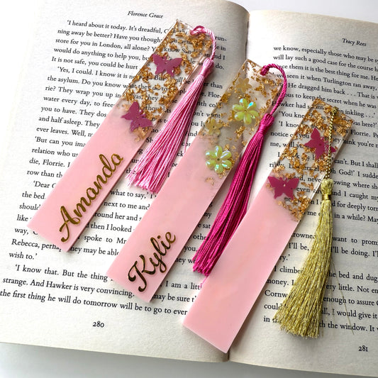 Three handmade resin bookmarks on an open book: personalised pink butterfly bookmark, personalised pink flower bookmark, and non-personalised pink butterfly bookmark, each with a matching tassel.