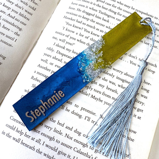 Personalised resin bookmark with blue, green, and silver foil in an open book. Handmade celestial-inspired bookmark for book lovers and readers.