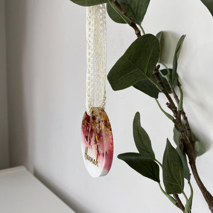 Personalised resin Christmas ornament with dried rose petals and gold leaf, angled to show thickness, custom name in gold vinyl, pink and white colours, lace ribbon included.