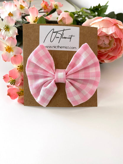 Single handmade pink cotton hair bow clip with gingham pattern on alligator clip displayed on branded packaging card, flowers in the background