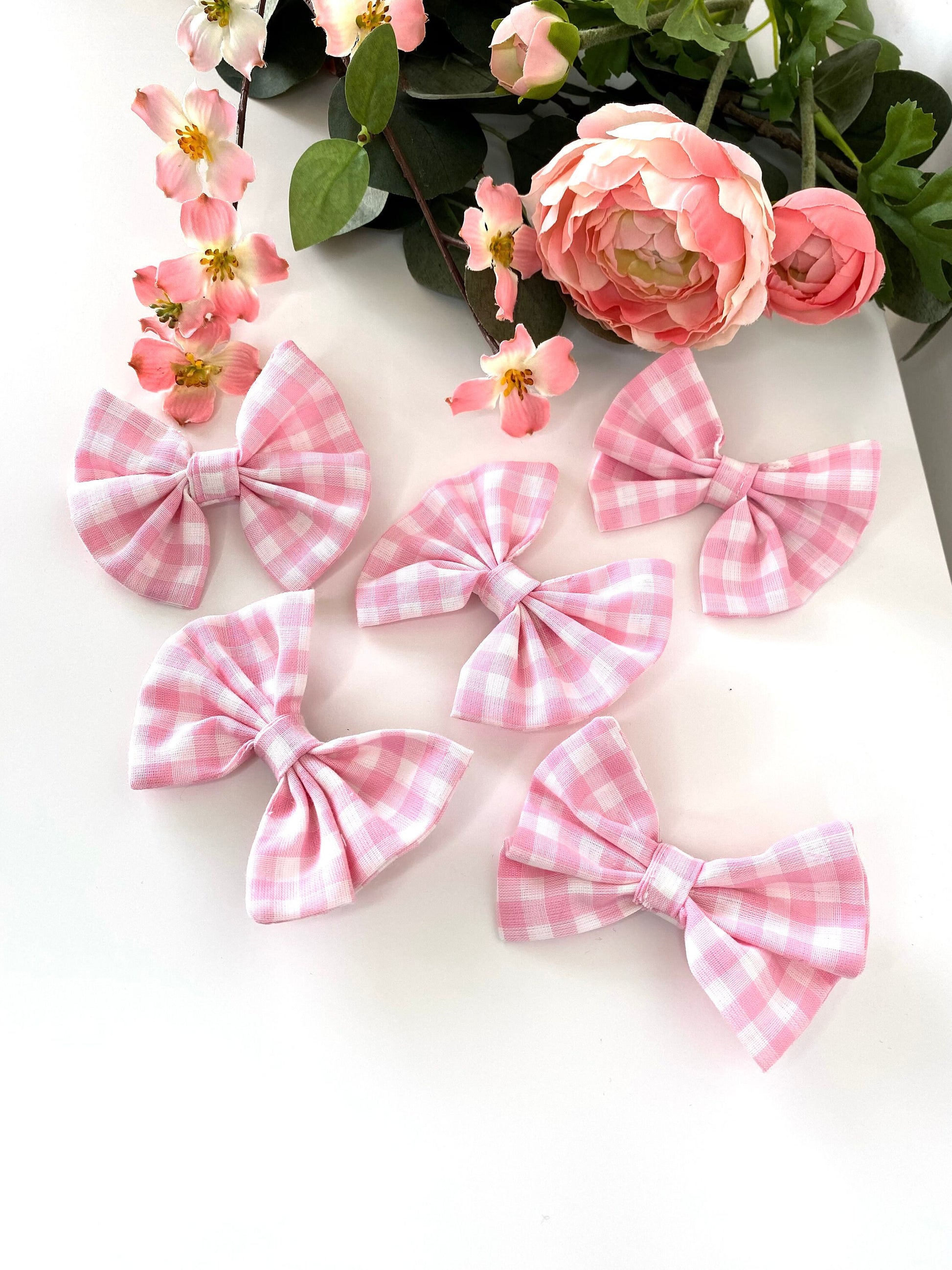 Five handmade pink cotton hair bow clips with gingham pattern on alligator clips displayed on a white surface with flowers, showing multiple individual clips