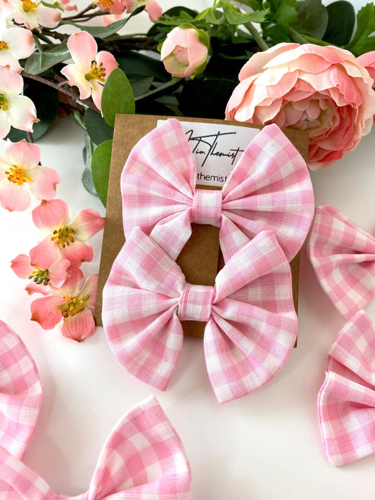 Two handmade pink cotton hair bow clips with gingham pattern on alligator clips displayed on branded packaging card, in front of flowers on a white surface