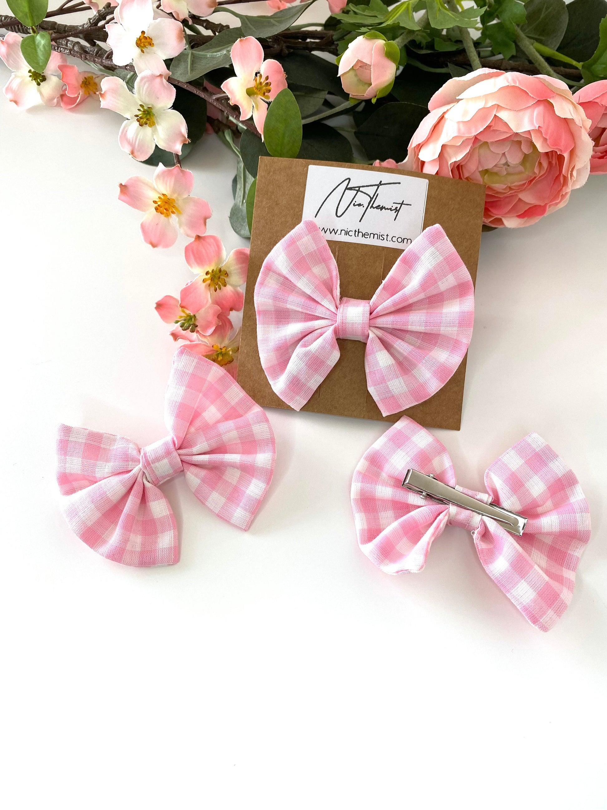 Single handmade pink cotton hair bow clip on branded packaging card with two additional clips scattered on white surface, one showing the gingham checkered bow and one showing the silver alligator clip, flowers in the background