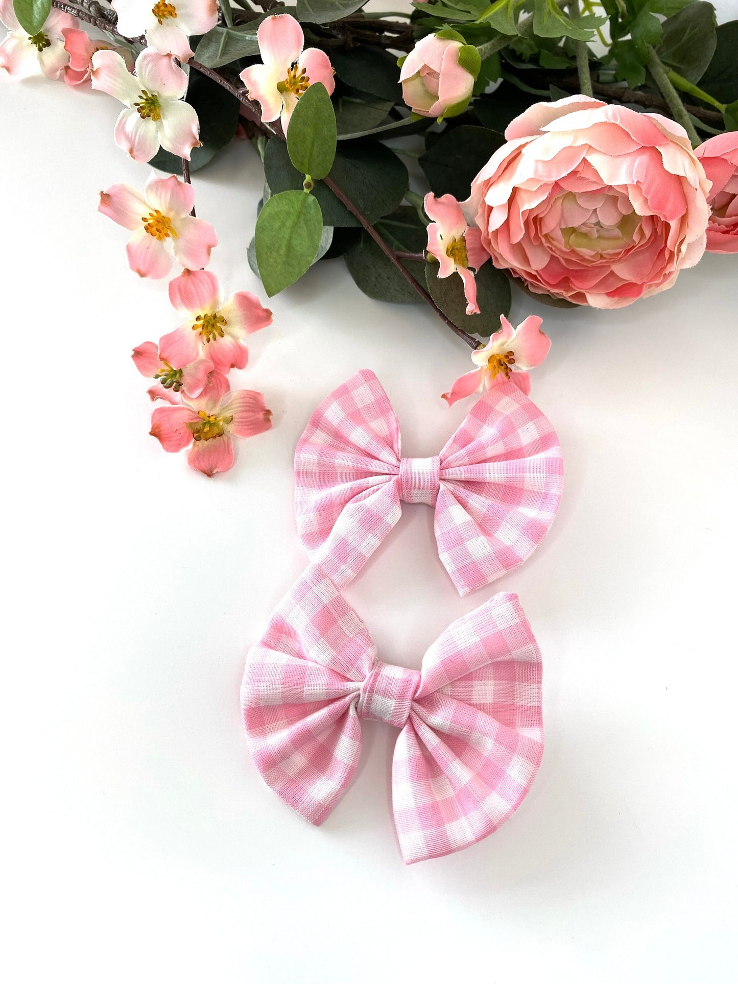 Two pink gingham hair bows on a white background with pink flowers in the background