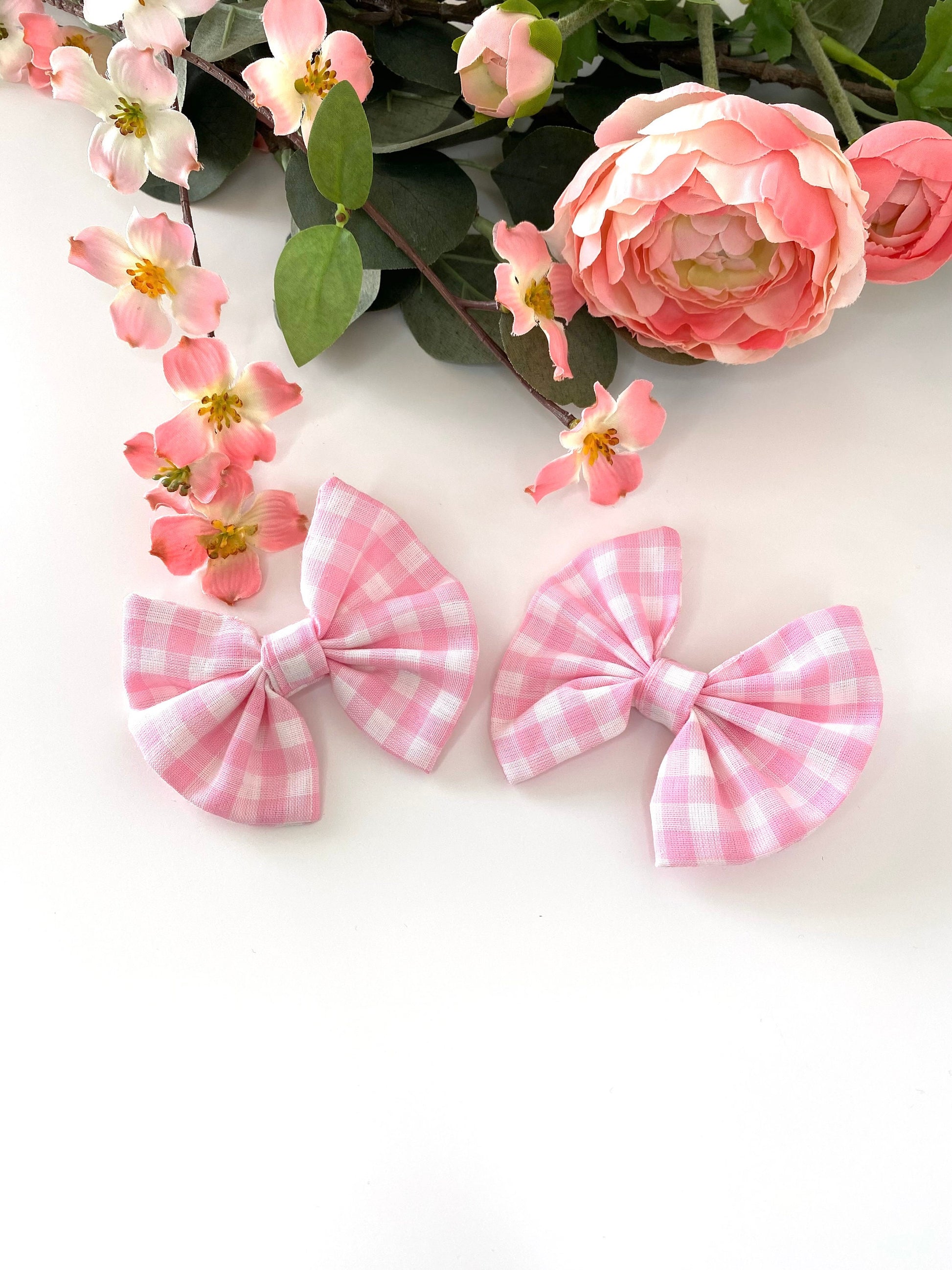 Two handmade pink and white cotton hair bow clips with gingham pattern on alligator clips, displayed facing up on a white surface with flowers in the background