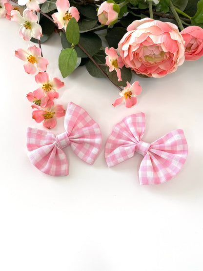 Two handmade pink and white cotton hair bow clips with gingham pattern on alligator clips, displayed facing up on a white surface with flowers in the background