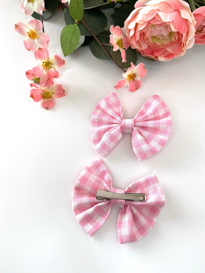 Two handmade pink cotton hair bow clips on a white surface with flowers in the background, one showing the gingham bow and the other showing the silver alligator clip
