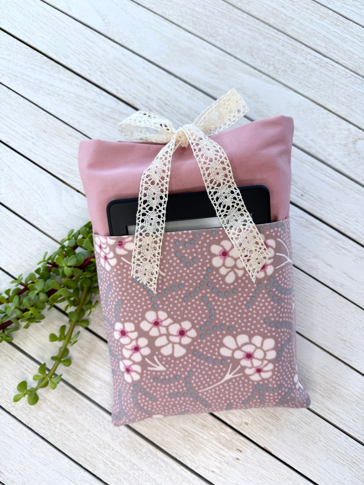 Handmade dusty pink book sleeve with a floral front pocket holding an e-reader, finished with a cream lace bow closure.