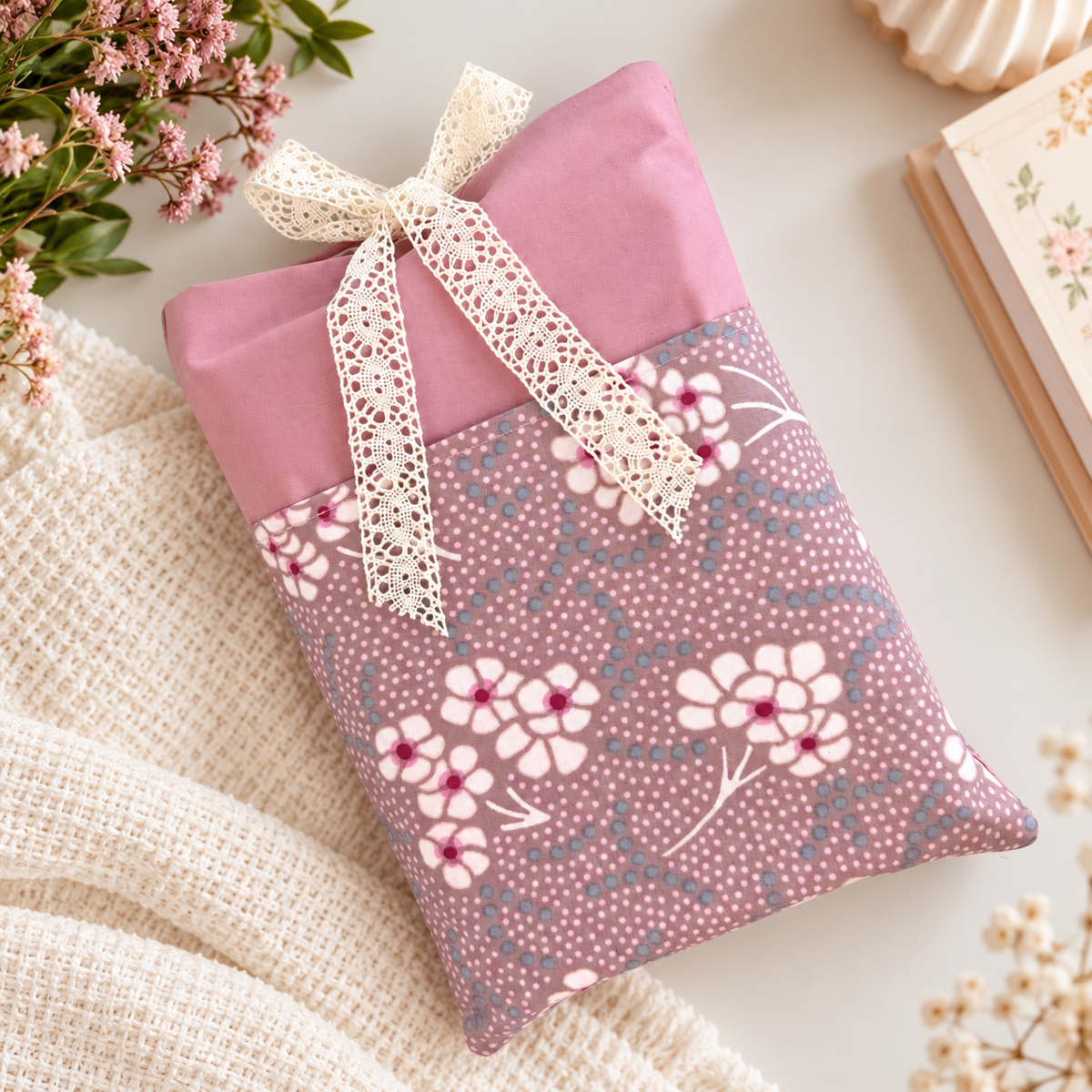 Handmade dusty pink fabric book sleeve with a pink and purple floral front pocket and cream lace bow closure, displayed beside flowers and a book.