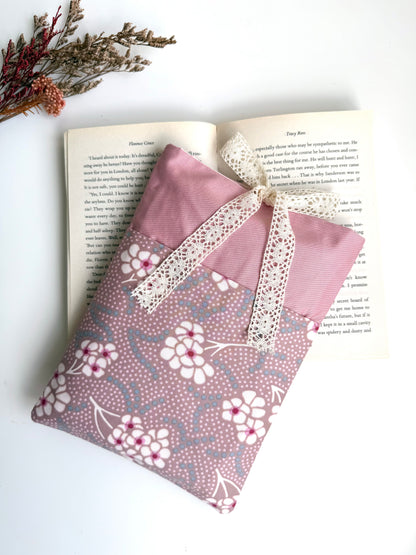 Handmade dusty pink fabric book sleeve with a floral front pocket and cream lace bow closure, displayed on an open paperback book.