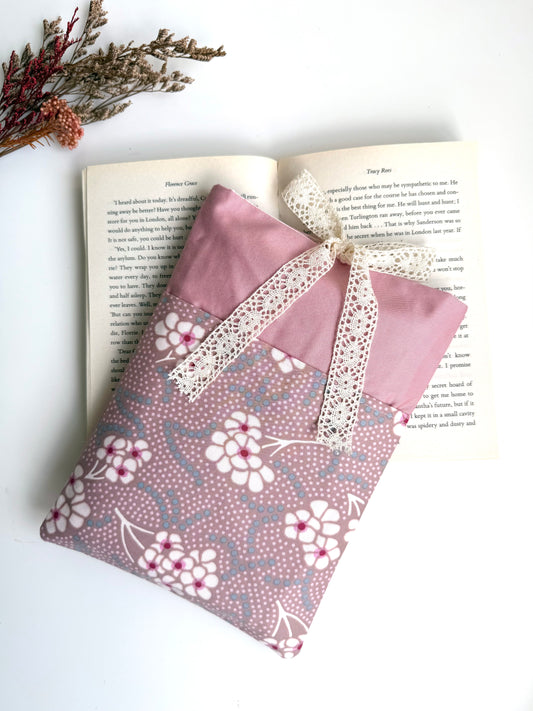 Handmade dusty pink fabric book sleeve with a floral front pocket and cream lace bow closure, displayed on an open paperback book.