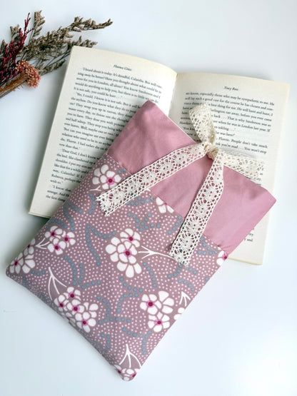 A pink fabric pouch with a white floral pattern and a cream lace ribbon bow, styled on top of an open book with dried flowers in the corner.
