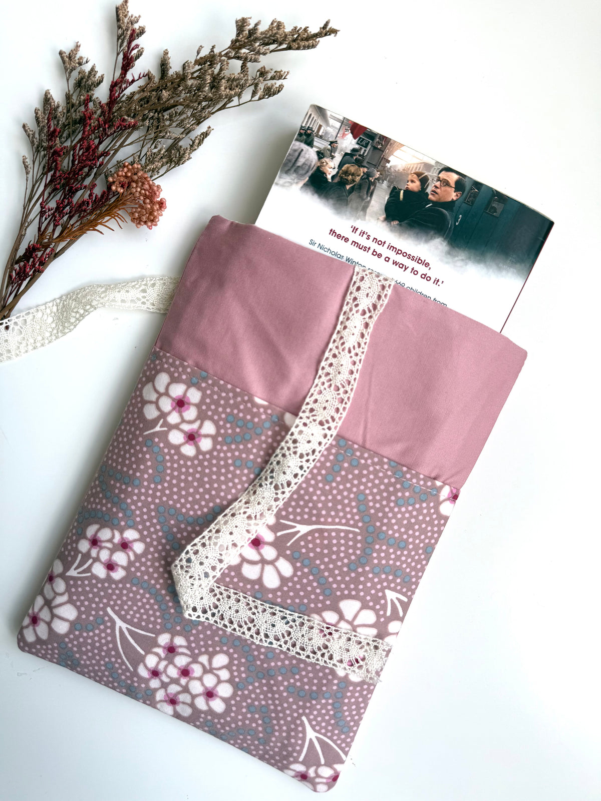 Handmade dusty pink floral book sleeve with a paperback book partially inserted, showing the fabric book cover, floral front pocket and cream lace ribbon closure.