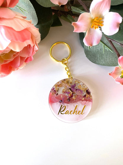 Pink and white round resin keychain with dried flowers and gold leaf, personalised in lowercase gold vinyl font, handmade resin keyring for gifts.