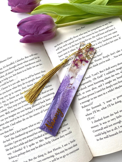 Purple resin bookmark with dried roses and gold leaf, personalised with gold vinyl, placed inside an open book, handmade bookmark for readers or gifts.