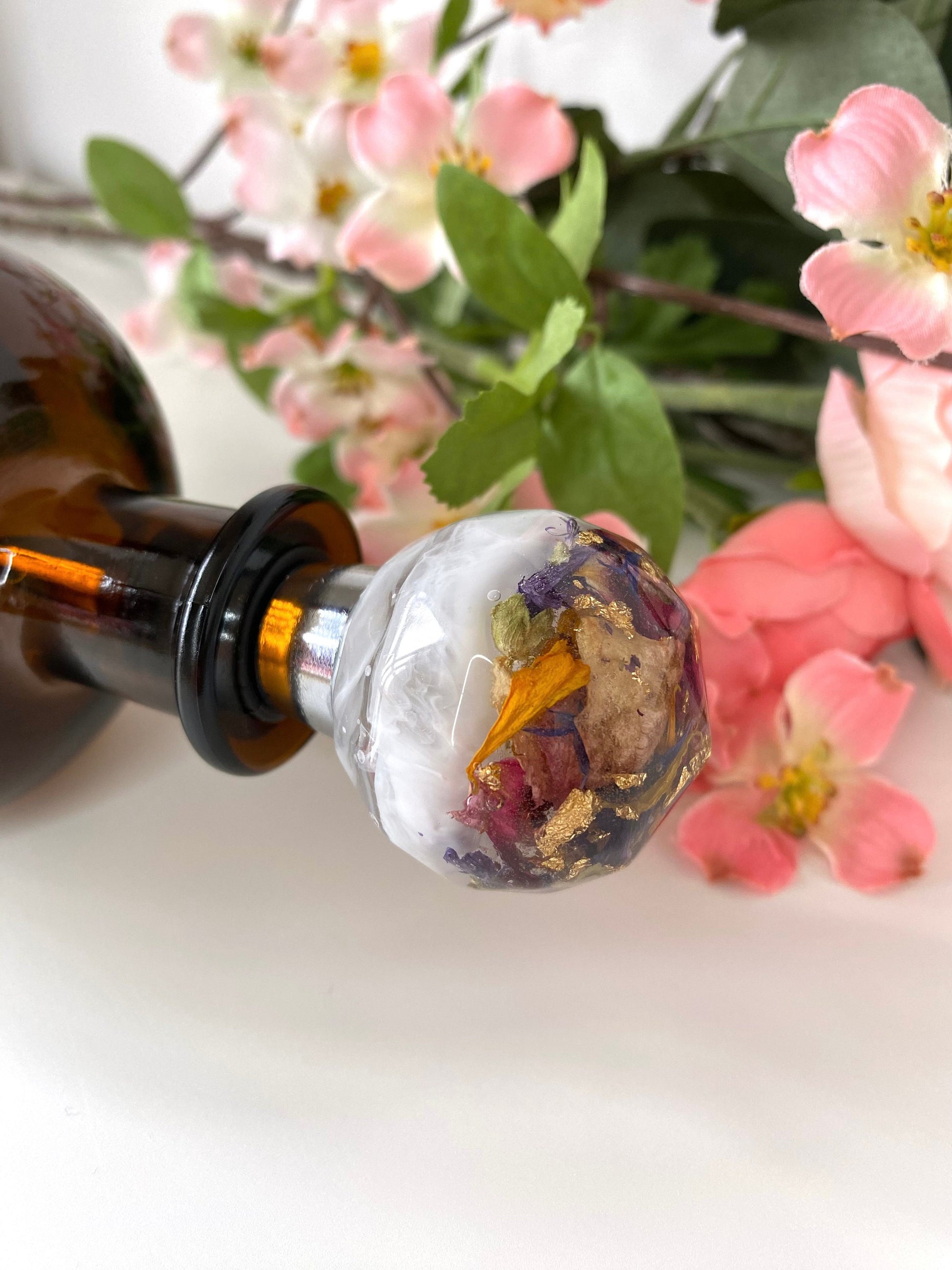 Close-up of handmade resin bottle stopper with stainless steel base, white resin top with clear resin containing colourful dried flowers and gold leaf, inserted into the mouth of a bottle lying flat on table, decorative and functional wine accessory for gifts or home use.