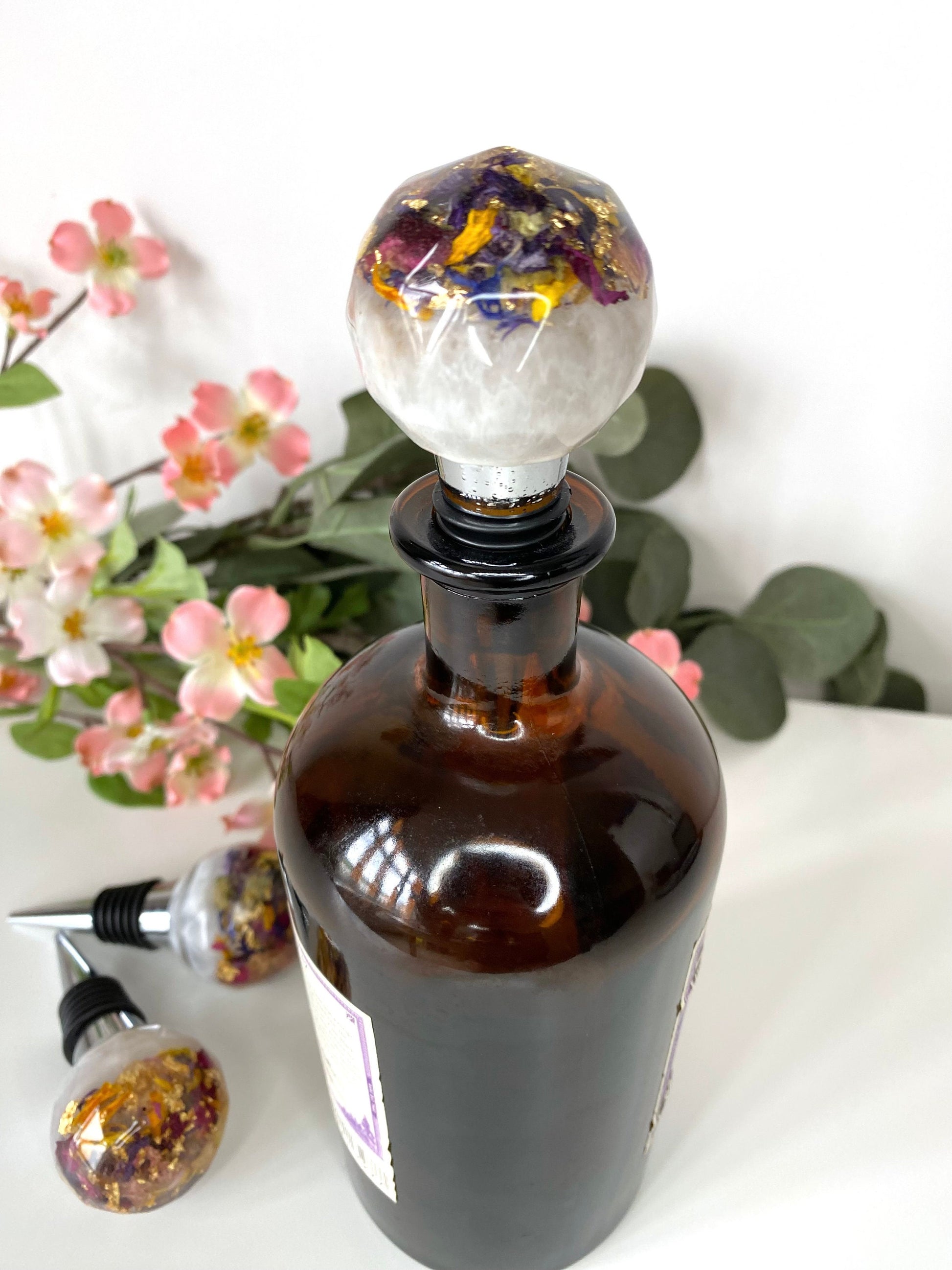 Handmade resin bottle stopper with stainless steel base, white resin top with clear resin containing colourful dried flowers and gold leaf, inserted into a bottle on a table, decorative and functional wine accessory for gifts or home use.