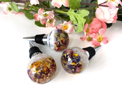 Close-up of three handmade resin bottle stoppers with stainless steel bases, white resin tops with clear resin containing colourful dried flowers and gold leaf, decorative and functional wine accessories for gifts or home use