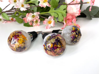 Three handmade resin bottle stoppers with stainless steel bases, white resin tops with clear resin containing colourful dried flowers and gold leaf, displayed in front of flowers, showing one full stopper and two focusing on resin tops, decorative and functional wine accessories for gifts or home use