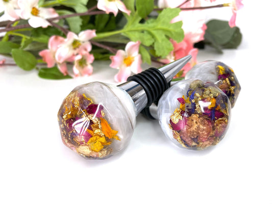Three handmade resin bottle stoppers with stainless steel bases, white resin tops with clear resin containing colourful dried flowers and gold leaf, displayed in front of flowers, showing one full stopper and two focusing on resin tops, decorative and functional wine accessories for gifts or home use.