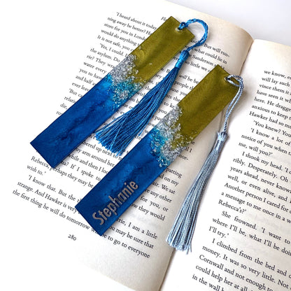 Two handmade resin bookmarks in an open book: one personalised with silver vinyl name, one plain. Blue, green, and silver celestial-inspired design, perfect gift for book lovers.