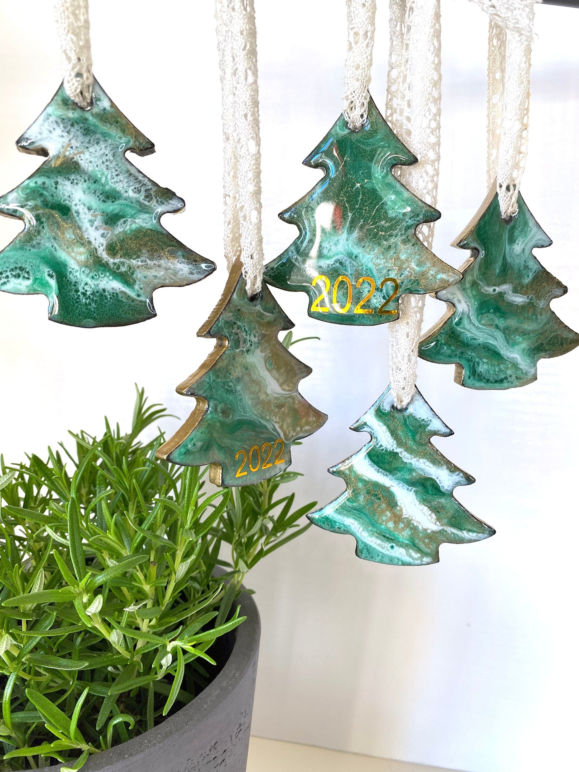 Close-up of tree-shaped resin Christmas ornaments on slate, highlighting unique resin design in green, white, and bronze/gold. Each ornament is one-of-a-kind with gold-painted edges and beige lace ribbon.