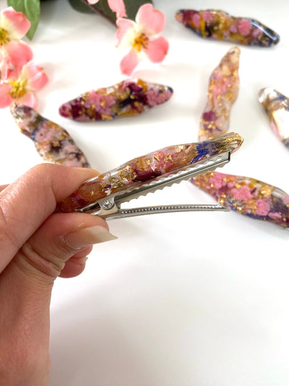 Hand holding a handmade resin alligator hair clip with dried flowers and gold leaf, showing the open clip mechanism and resin design