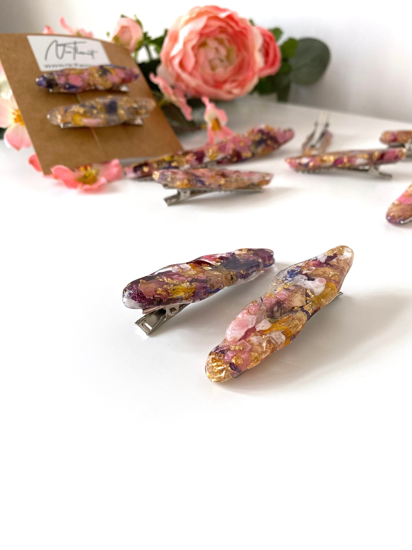 Close-up of two handmade resin alligator hair clips with dried flowers and gold leaf in sharp focus, with packaging and scattered clips blurred in the background