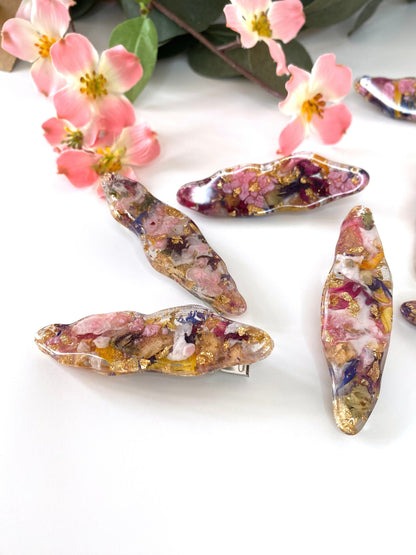 four handmade resin alligator hair clips with dried flowers and gold leaf, scattered on a surface with flowers in the background, each clip uniquely designed
