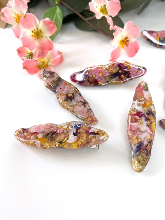 four handmade resin alligator hair clips with dried flowers and gold leaf, scattered on a surface with flowers in the background, each clip uniquely designed
