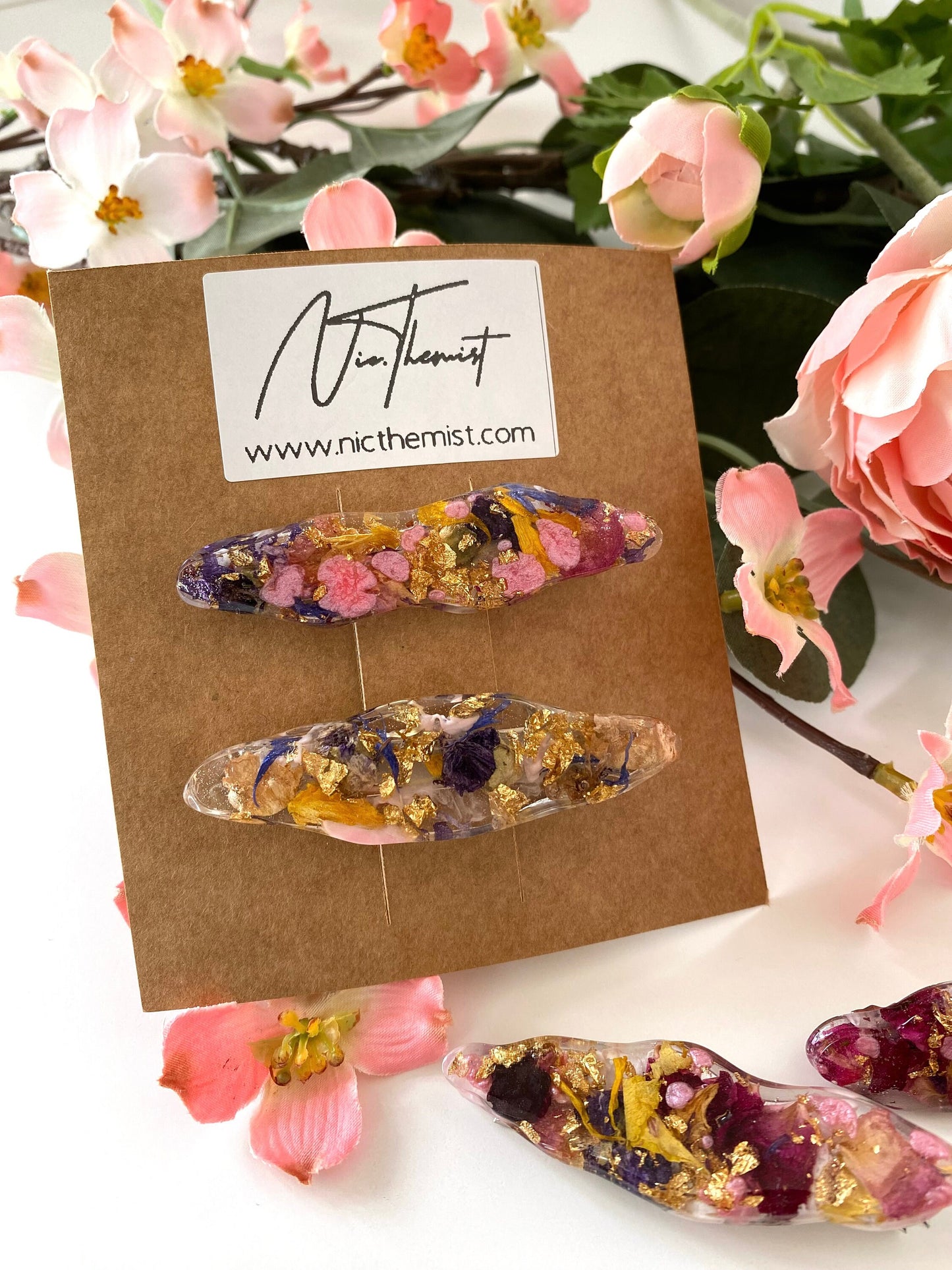 Close-up of two handmade resin alligator hair clips on branded packaging card with logo, showing dried flowers and gold leaf details
