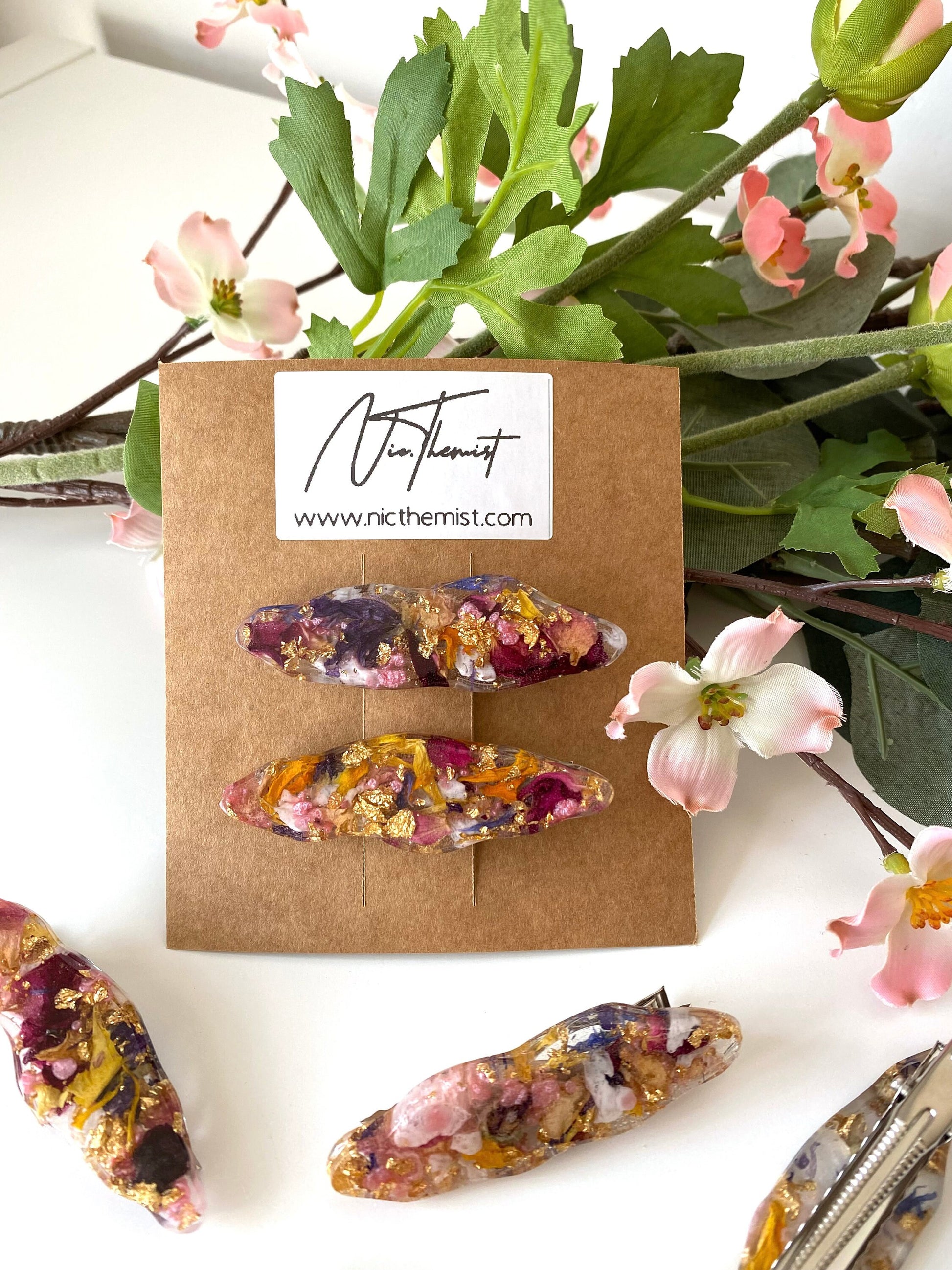 Close-up of handmade resin alligator hair clips with dried flowers and gold leaf on branded packaging card, surrounded by additional clips scattered on a white surface